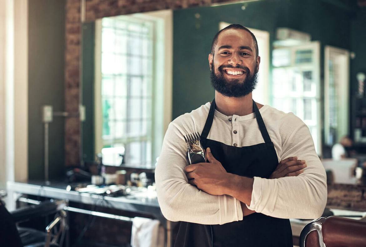 barbeiro sorrindo para a câmera com a bancada de uma barbearia ao fundo
