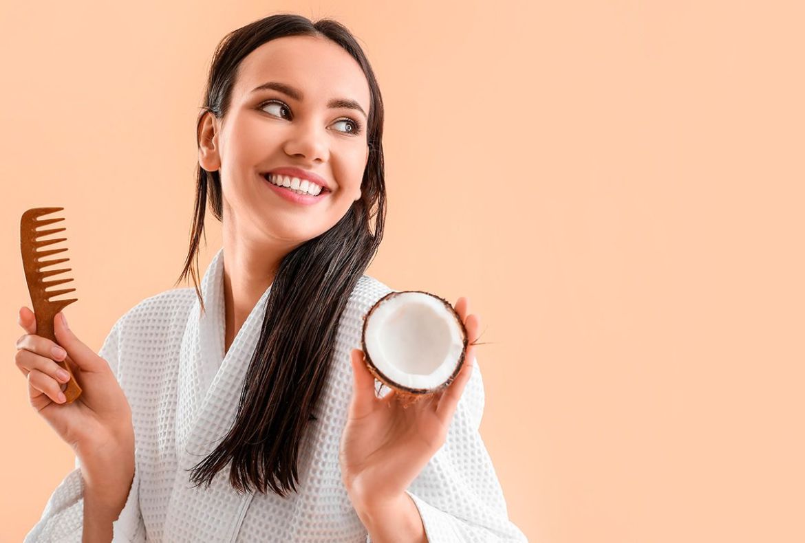 mulher segurando um pente e um coco seco aberto. ela usa roupão e tem óleo de coco no cabelo